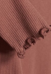 Rippstrickstoff in einem sanften Rosaton mit texturierten, fransigen Kanten. Das Material zeigt eine weiche, leichte Dehnung und einen subtilen Glanz.