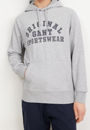 Osoba ubrana w jasnoszarą bluzę z kapturem z napisem "Original Gant Sportswear", ciemne spodnie, jedna ręka w przedniej kieszeni, na jednolitym tle.