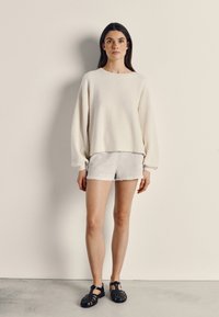 Kremo spalvos vilnos džemperis su balioninėmis rankovėmis, derinamas su pilkomis šortais. Modelis dėvi juodus dirželinius sandalus. Neutralios spalvos fonas.