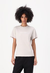 Tricou bej deschis cu mâneci scurte și guler rotund. Prezintă un logo alb "Calvin Klein" centrat pe piept. Material moale.