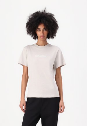 Helles beiges T-Shirt mit kurzen Ärmeln und Rundhalsausschnitt. Mit einem weißen "Calvin Klein"-Logo, das auf der Brust zentriert ist. Weiches Material.