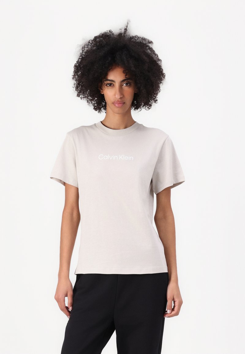 Tricou bej deschis cu mâneci scurte și guler rotund. Prezintă un logo alb "Calvin Klein" centrat pe piept. Material moale.
