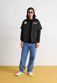 Black short-sleeve shirt with two front pockets and logo patches, paired with blue loose-fit jeans and white sneakers.圆形太阳镜。