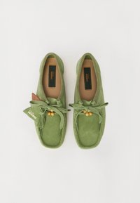 Des chaussures slip-on en suède vert avec un bout rond, des lacets et des perles en bois décoratives. Comprend une étiquette marron et une semelle texturée.