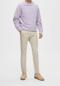 Sweatshirt violet clair avec des manches raglan, pantalon beige slim-fit, et chaussures beige avec une semelle en caoutchouc, posant devant un fond blanc.