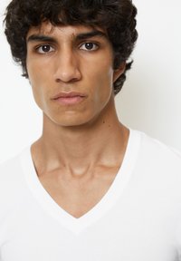 Witte v-hals t-shirt op een persoon met krullend donker haar en een neutrale uitdrukking. Kenmerken zijn een gladde textuur en een aansluitend ontwerp.
