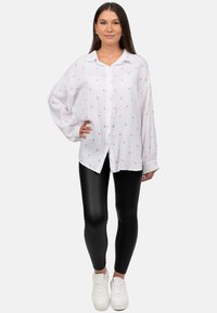 Witte blouse met een roze hartpatroon, lange mouwen en een relaxte pasvorm, gecombineerd met zwarte leggings en witte sneakers.