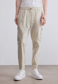 Μπεζ cargo παντελόνια με ελαστική μέση, κορδόνι ρύθμισης και πλαϊνές τσέπες, σε συνδυασμό με λευκά αθλητικά παπούτσια σε λείο επιφάνεια.