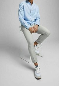 Světle modrá košile na knoflíky s kapsou, šedé kalhoty a bílé tenisky s šedými a béžovými akcenty, sedící na bílé židli.