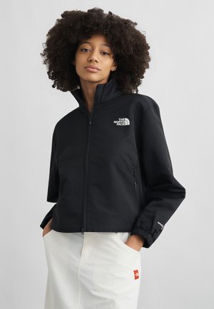 Γυναίκα με σγουρά μαλλιά που φοράει μαύρο μπουφάν North Face με φερμουάρ και λευκή φούστα, με τα χέρια στις τσέπες, ουδέτερο φόντο.