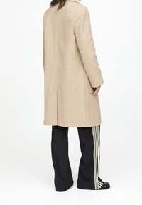 Abrigo de lana beige con corte recto, dos bolsillos laterales y una abertura en la espalda, combinado con pantalones negros con rayas verticales blancas.