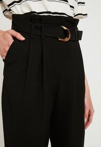 Černé kalhoty s vysokým pasem a záhyby, vybavené sladěným páskem s kulatou hnědou sponou. Měkká látka, střih na míru.