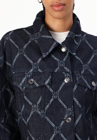 Donkerblauwe denimjack met contrasterend lichtblauw geometrisch patroon. Voorzien van zilveren knopen en borstzakken met kleppen.