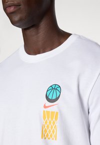 Bela bombažna majica s grafičnim dizajnom, ki prikazuje teal košarko in rumeno košarkarsko mrežo, okrašena z rdečim logotipom Nike.