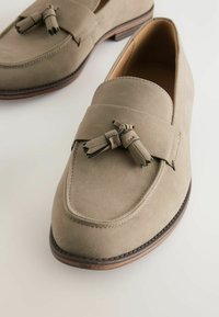 Mocassins en suede beige avec une semelle plate et des accents en bois, ornés de franges décoratives à l'avant et de coutures le long des bordures.
