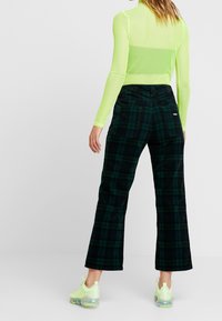 Femme portant un haut transparent à manches longues vert fluo, un pantalon à carreaux vert foncé et noir, et des baskets vert fluo, se tenant devant un fond uni.