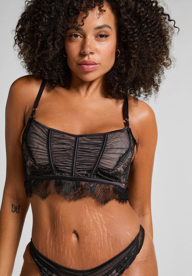 Mujer con cabello rizado vistiendo lencería de encaje negra, mostrando estrías en su abdomen y un tatuaje de mariposa en su brazo.
