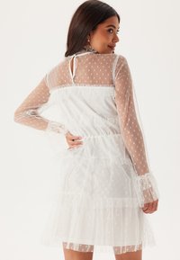 Vit klänning med genomskinligt tyg med prickar, volanglager och långa ärmar. Har en nyckelhålsrygg och elastisk midja. Mjuk textur.