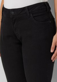 Zwarte high-waist jeans met knoop en rits, gedragen met een witte top, waarbij de voorzakken zichtbaar zijn en een deel van de rechterhand op de dij rust.