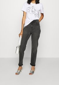 Donna che indossa una t-shirt bianca con design di faccia astratta, jeans neri arrotolati, sandali con tacco argento, mentre tiene una borsa bianca con texture.