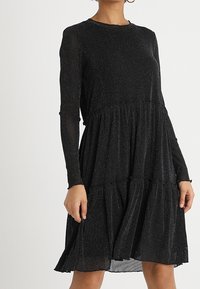 Black long-sleeve dress with a tiered hem, made of sparkly fabric. Features a round neckline and a relaxed fit.