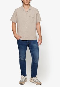 Short-sleeve, light beige shirt made of textured fabric, featuring a button front and pocket. Paired with blue jeans and gray sneakers.