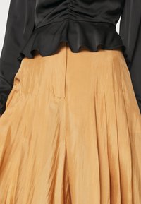 Haut noir à manches longues avec détail froncé et ourlet à volants, associé à un pantalon large taille haute, plissé, de couleur marron clair.