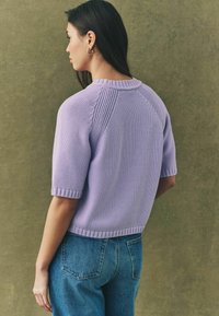 Maglione corto a maniche corte lilla con collo e polsini a coste, abbinato a jeans blu a vita alta. Tessuto testurizzato.