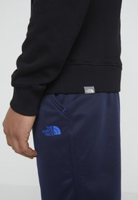 Felpa nera con polsini a costine, abbinata a pantaloni jogger blu con logo e cuciture a contrasto sui taschini. Tessuto morbido, vestibilità casual.