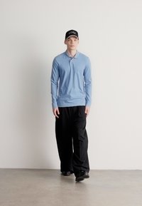 Šviesiai mėlyna ilgomis rankovėmis polo marškinėliai, juodos krovinių kelnės ir juoda kepuraitė. Lygus audinys, laisvas kirpimas ir minimalus prekės ženklo ženklinimas marškinėliuose.