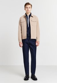 Giacca beige con due tasche anteriori, indossata sopra una camicia button-up scura e pantaloni in denim scuri. Stivali neri alla caviglia completano l'outfit.