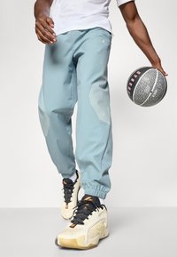 Himmelblå joggebukser med elastiske mansjetter, med prikkete mønster på utsiden av lårene, parret med svart og kremfargede basketballsko og en ball.