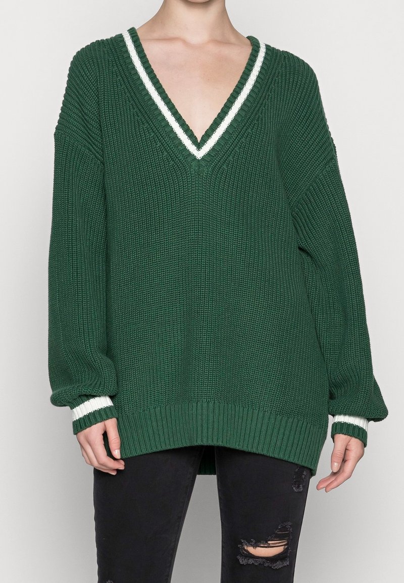 Pull en maille vert foncé à manches longues avec un col en V profond et un liseré blanc, porté avec un jean noir déchiré.