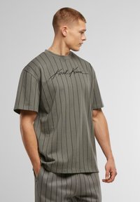 Camiseta verde oliva con rayas verticales negras, escote redondo, mangas cortas y "Karl Kani" estampado en cursiva.