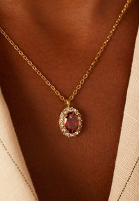 Gouden ketting met een ovale rode edelsteen in het midden, omringd door kleine heldere kristallen, rustend op een gebruinde huid.