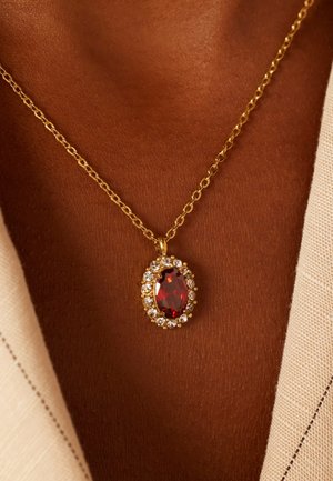 Gouden ketting met een ovale rode edelsteen in het midden, omringd door kleine heldere kristallen, rustend op een gebruinde huid.