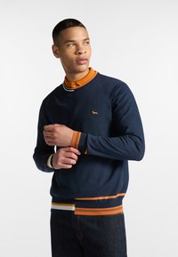 Námořnická crewneck svetr s oranžovými, žlutými a bílými pruhovanými akcenty na manžetách a lemu, který má na hrudi malé vyšívané logo.