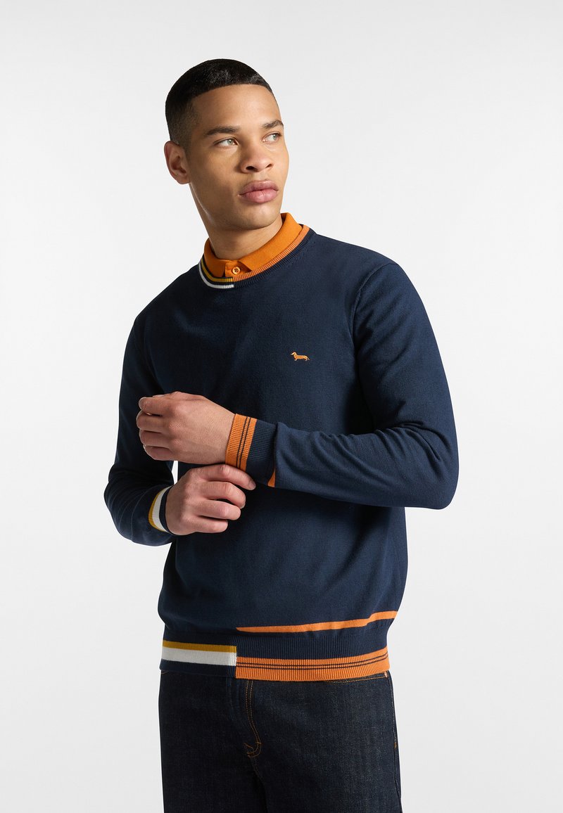 Námořnická crewneck svetr s oranžovými, žlutými a bílými pruhovanými akcenty na manžetách a lemu, který má na hrudi malé vyšívané logo.