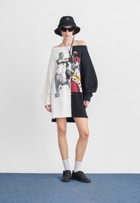 Άτομο που φοράει υπερμεγέθη φόρεμα με γραφικό μπάσκετ μισό λευκό, μισό μαύρο, μαύρο καπέλο τύπου bucket, γυαλιά ηλίου, λευκές κάλτσες και μαύρα αθλητικά παπούτσια σε μπλε πάτωμα.