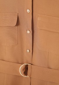 Camisa marrón con dos bolsillos en el pecho, botones dorados y un cinturón a juego con una hebilla metálica. Textura suave con patrones sutiles.