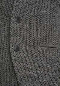 Szary teksturowany blezer z tkanym wzorem, wyposażony w dwa czarne guziki i boczną kieszeń, wykonany z miękkiej tkaniny.