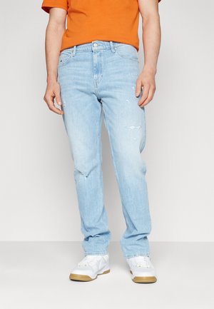 Mann trägt hellblaue, ausgewaschene Jeans, weiße Sneakers mit Gummisohlen und ein oranges Hemd und steht vor einem schlichten hellen Hintergrund.