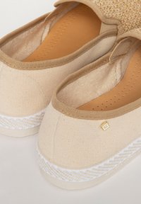 Beige canvas instapschoenen met een geaccentueerde textuurweving, een zachte voering aan de binnenkant en een witte gevlochten zool. Gouden logo detail aan de zijkant.