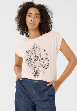 Haut rose clair à manches courtes avec un imprimé mandala noir détaillé. Le tissu est doux avec une texture lisse, associé à un pantalon en denim.