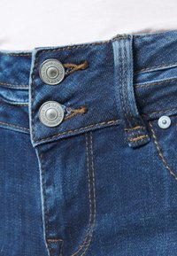 Mörkblå denimjeans med dubbel knappstängning, kontrasterande guldstickning och en slät textur, med synliga fickstickningsdetaljer.