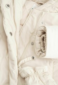 Kremowa kurtka puchowa z miękkim wnętrzem, z metalowymi zatrzaskami, elastycznymi mankietami i szczegółami w strukturalnym ściegu.