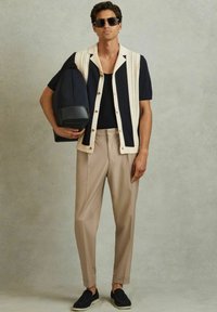 Cárdigan de punto azul marino y crema de manga corta, top negro, pantalones beige con pliegues y zapatos oscuros sin cordones, sosteniendo un bolso azul marino.