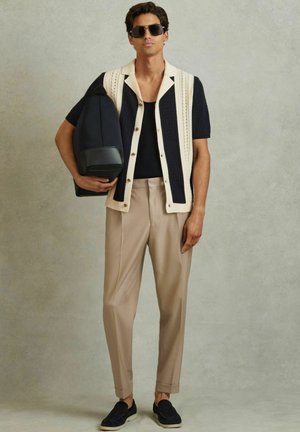 Navy and cream knitted short-sleeve cardigan, black top, beige pleated trousers, and dark slip-on shoes, holding a navy bag.