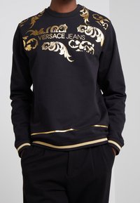 Sweatshirt noir en matériau doux avec des motifs dorés ornés et le texte "VERSACE JEANS". Comprend des rayures dorées à l'ourlet.