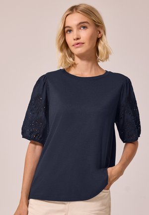Femme aux cheveux blonds courts portant un haut bleu marine avec des manches bouffantes brodées et un pantalon beige, debout devant un fond uni.